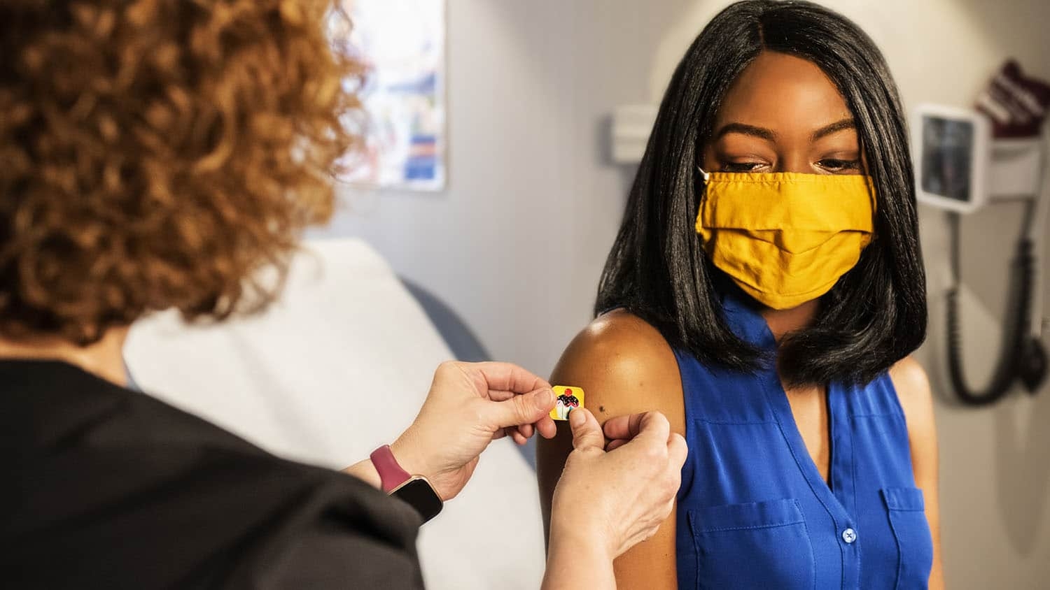 woman with red hair puts band-aid on black woman wearing a face mask; the image suggests that the black woman just received her vaccine shot