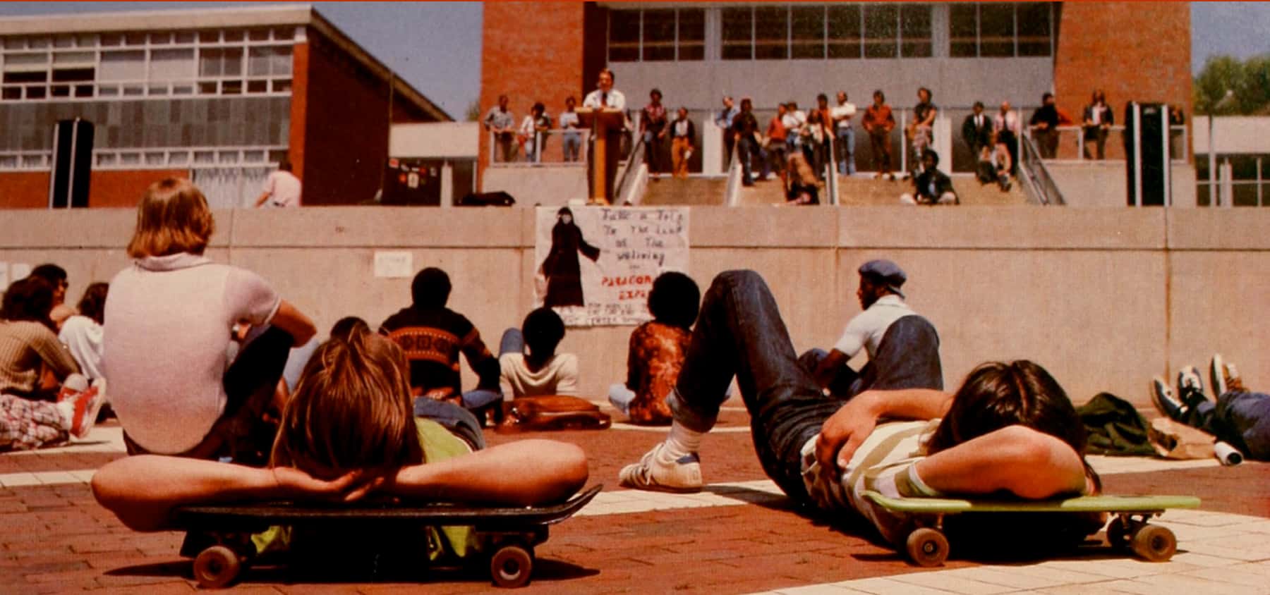 Students lie on skateboards in the brickyard while listening to a speaker