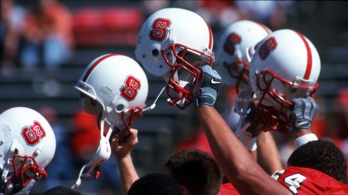 NC State football players raise their helmets in unity as they walk off the field.