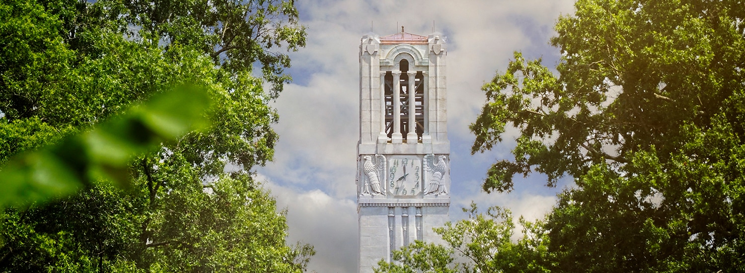 The top of NC State's Memorial Belltower peeking through leafy trees.