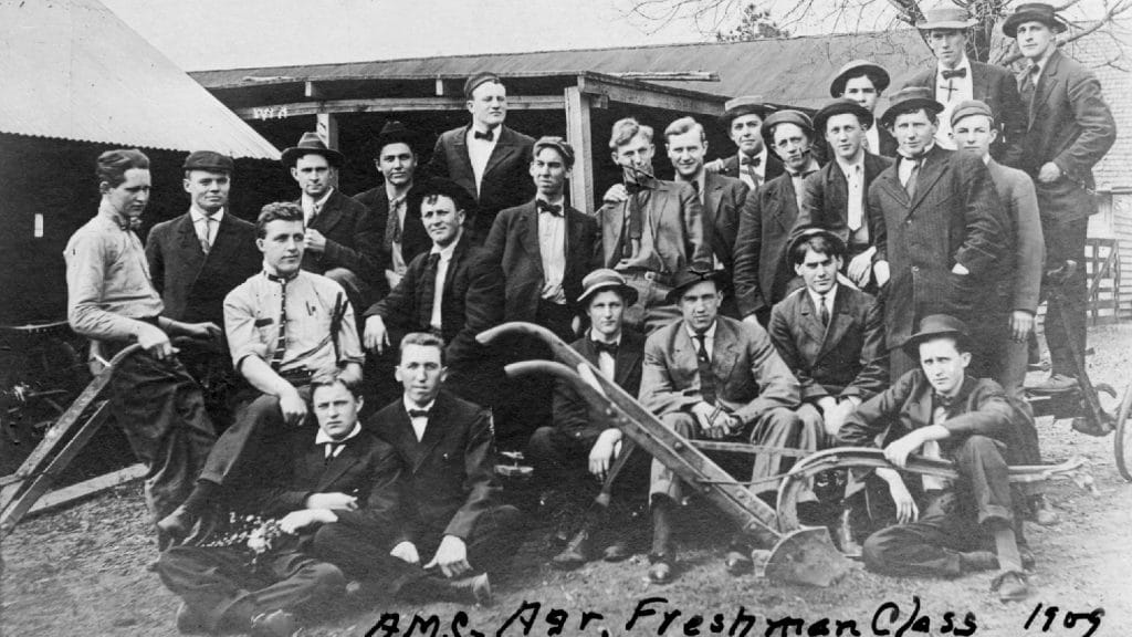 A black and white photo of a group of men wearing overalls, posing in an enginneering classroom..