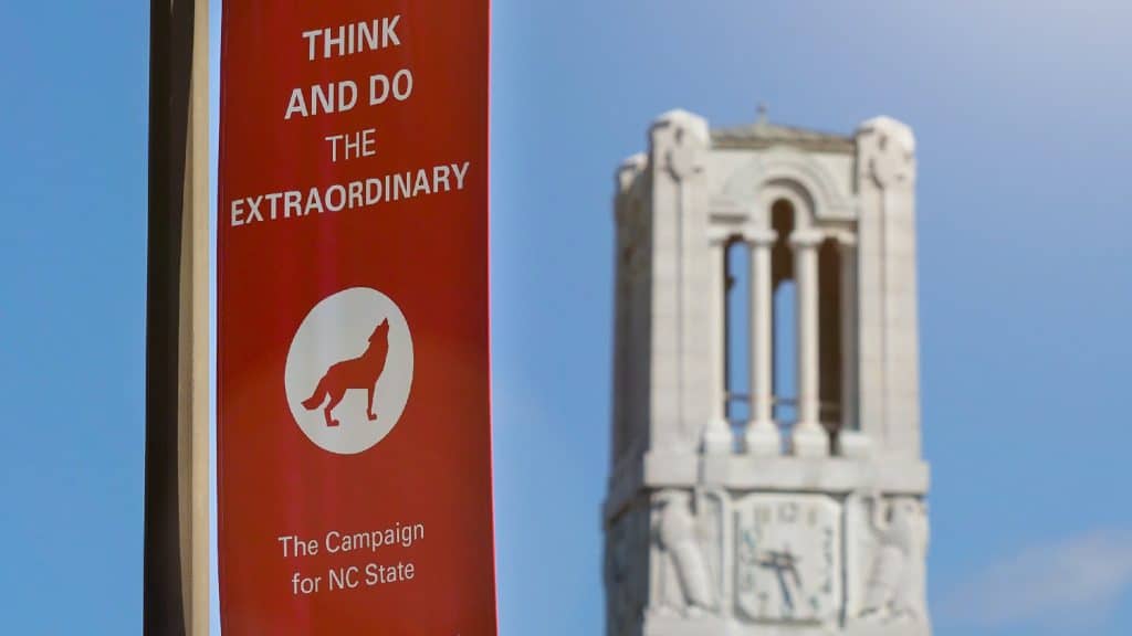 Hanging near the Memorial Belltower, a red banner with a wolf logo reads "Think and Do the Extraordinary: The Campaign for NC State."