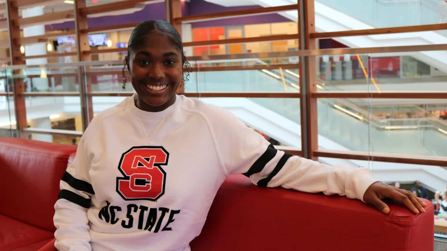 A young woman in a white NC State sweater sits in Talley Student Union.