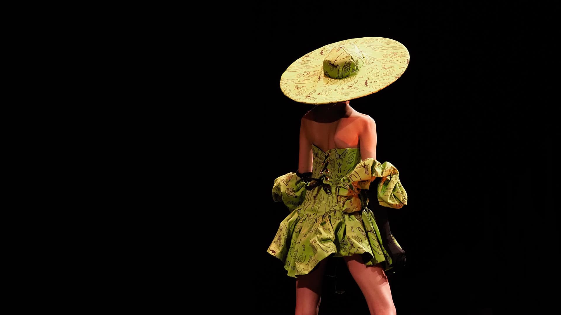 The back of a student walking down a runway wearing a modern green garment with a large hat.