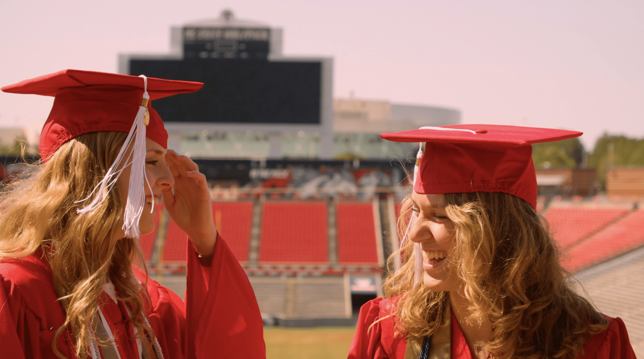 Students Alyssa McInnis and Emily Southard stand in red graduation robes in Carter-Finley Stadium.