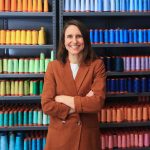Faculty member Kate Nartker stands in front of a wall of colorful spools of thread.