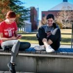 Two students chat and scroll through their smartphones in an outdoor courtyard.