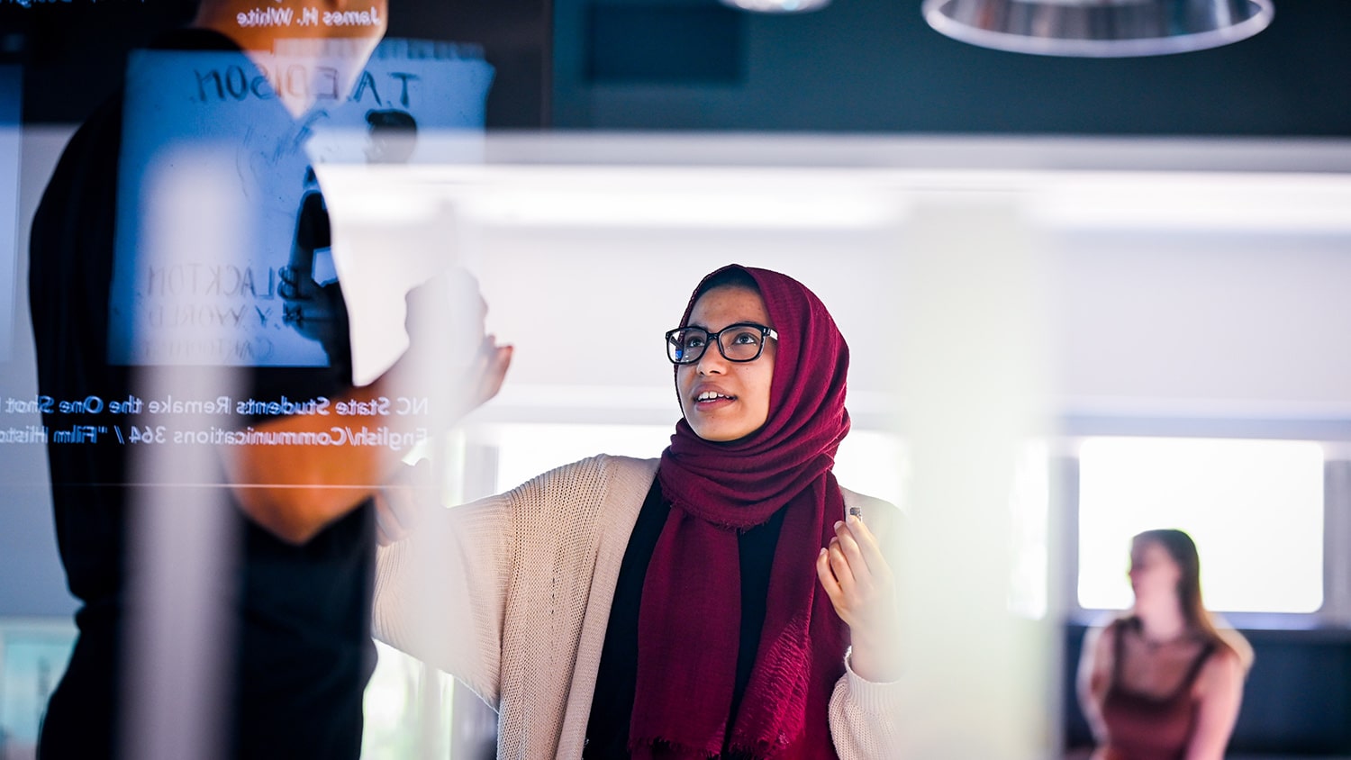 Image captured through a window with projections displayed on the surface, shows a female student gesturing while talking to someone in the Innovation Studio in D.H. Hill Library.