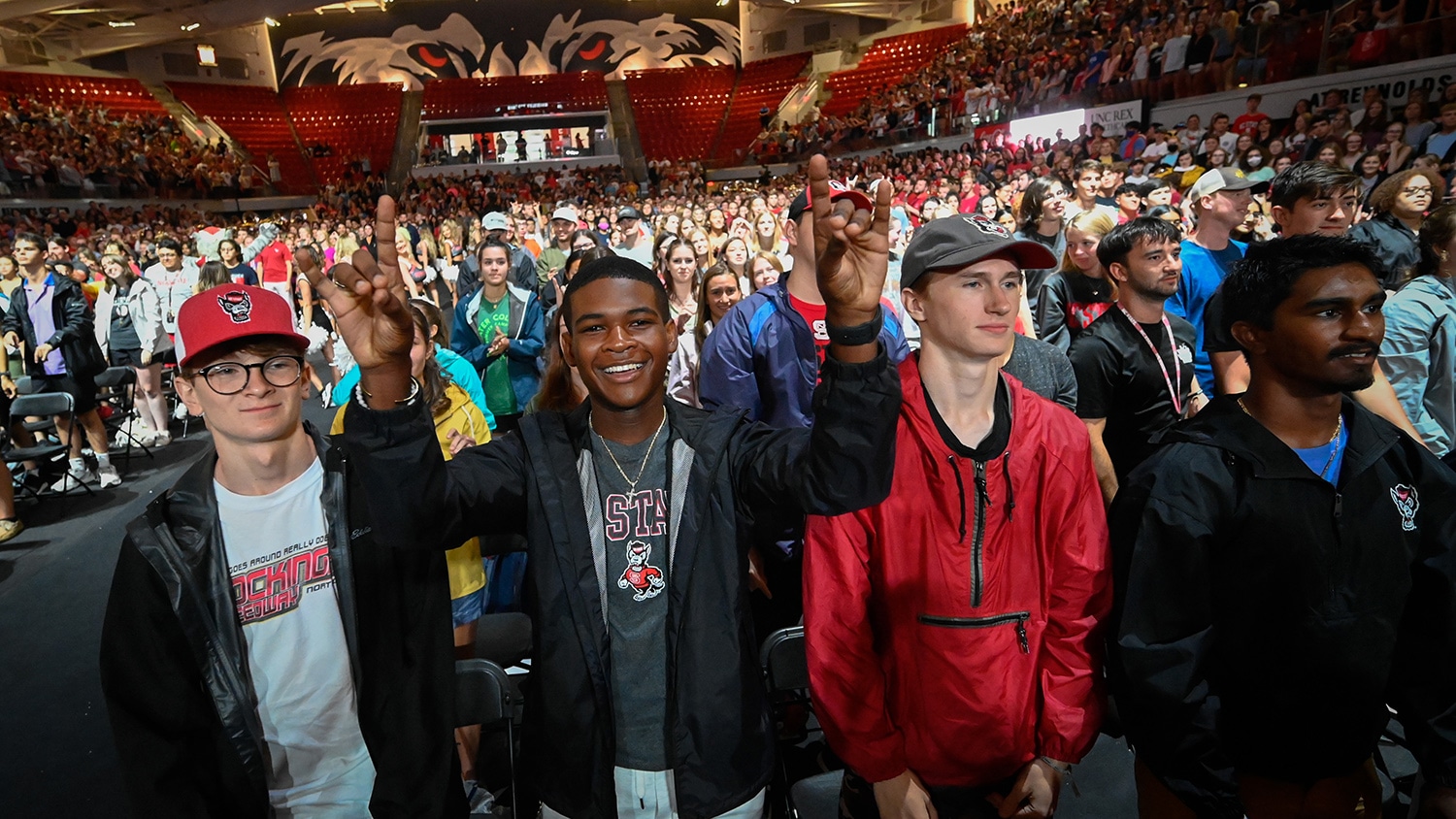 A student holds up his hands in the "wolfie" gesture while surrounded by other students in Reynolds Coliseum.