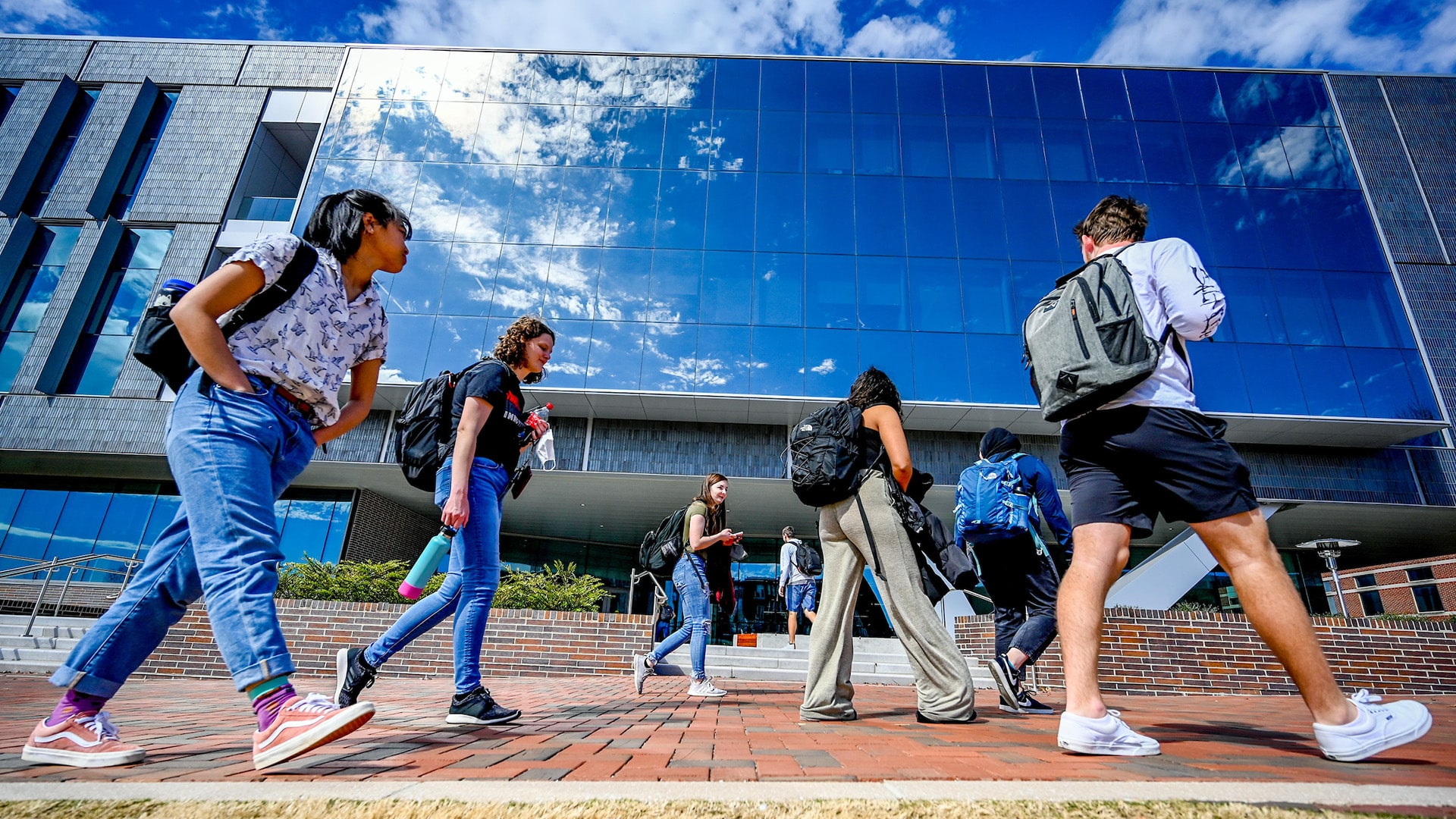 Students head for class on NC State's Centennial Campus, with the sky reflected in glass behind them.