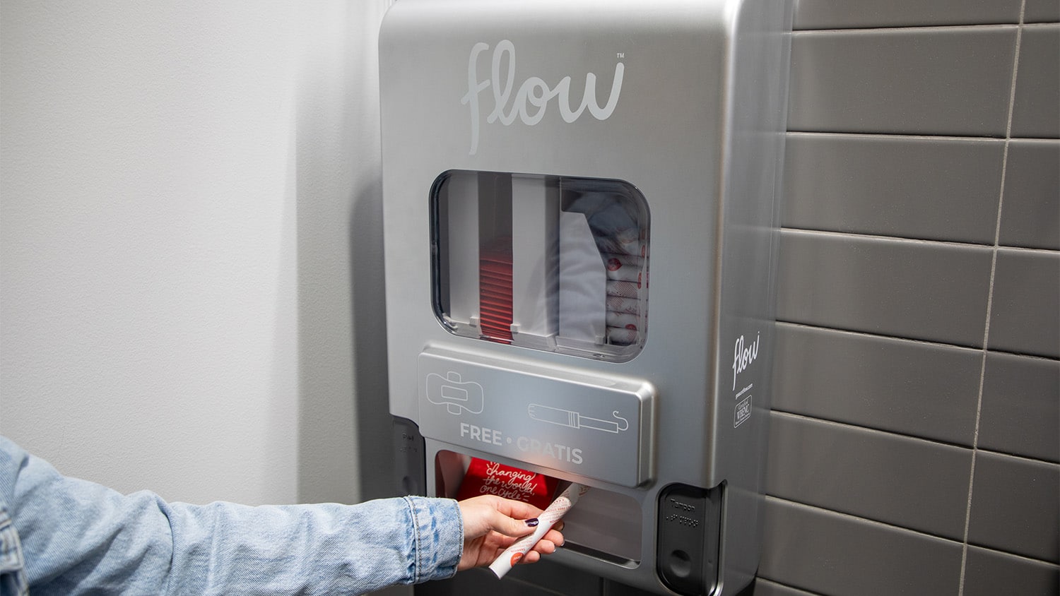 A hand pulls a tampon from a dispenser on a restroom wall.
