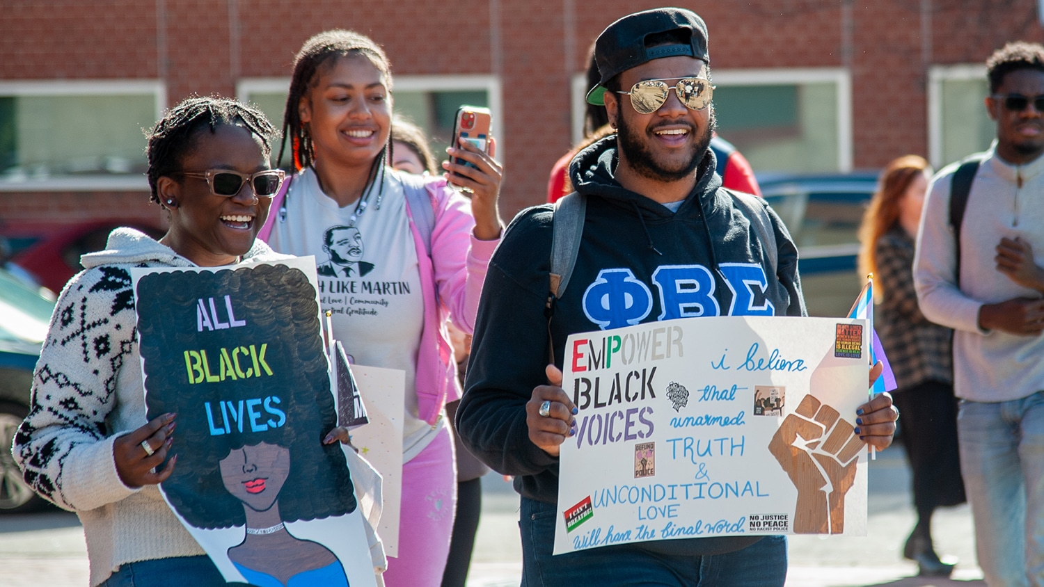 Three people hold handmade signs while walking at the "March Like Martin" event, commemorating Martin Luther King, Jr.