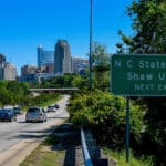 The downtown Raleigh skyline with a highway sign pointing to NC State's nearby campus.