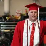 New graduate Isaac Mayle stands in the Pack Motorsports workshop in his red cap and gown.