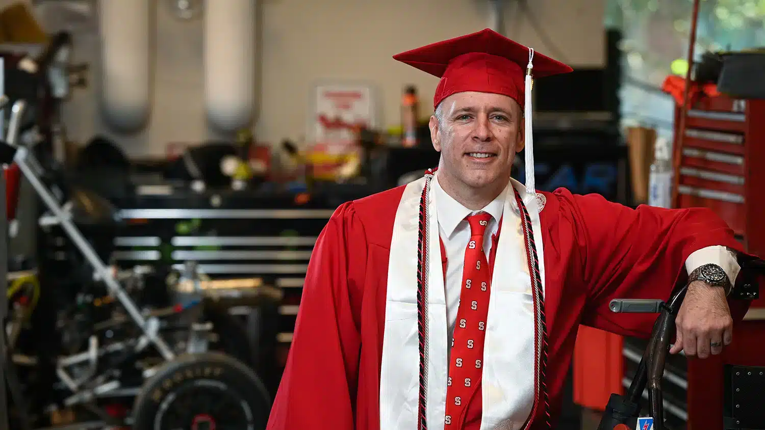 New graduate Isaac Mayle stands in the Pack Motorsports workshop in his red cap and gown.