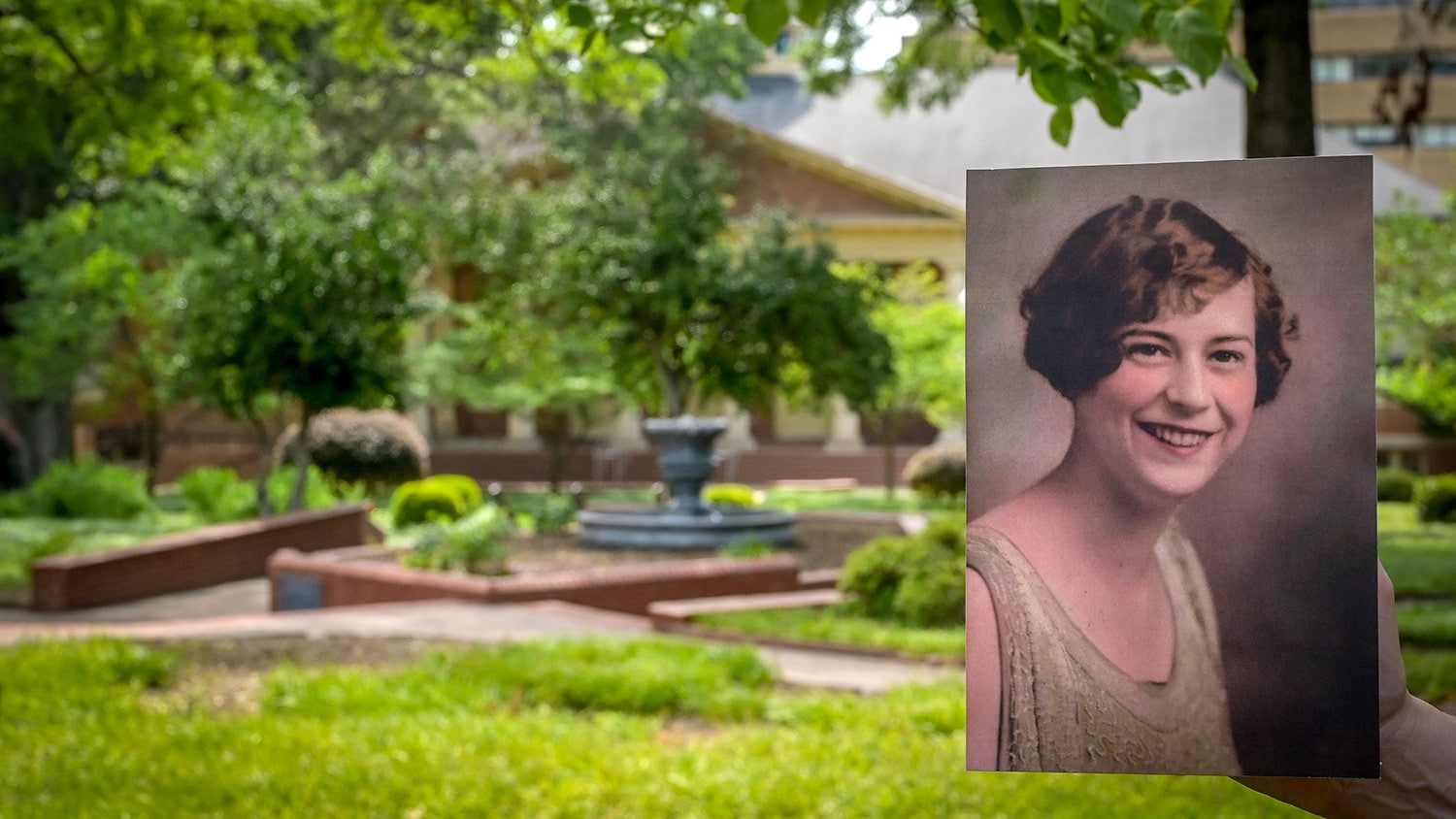 A colorized photo of a young adult Mary Yarbrough is held up in front of a swath of greenery, brick pathways and a fountain at Yarbrough Court.