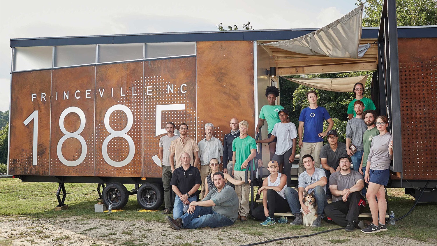 A group of students from the College of Design sit and stand in front of the Princeville mobile museum, which has the words "Princeville, N.C., 1885" printed on the side.