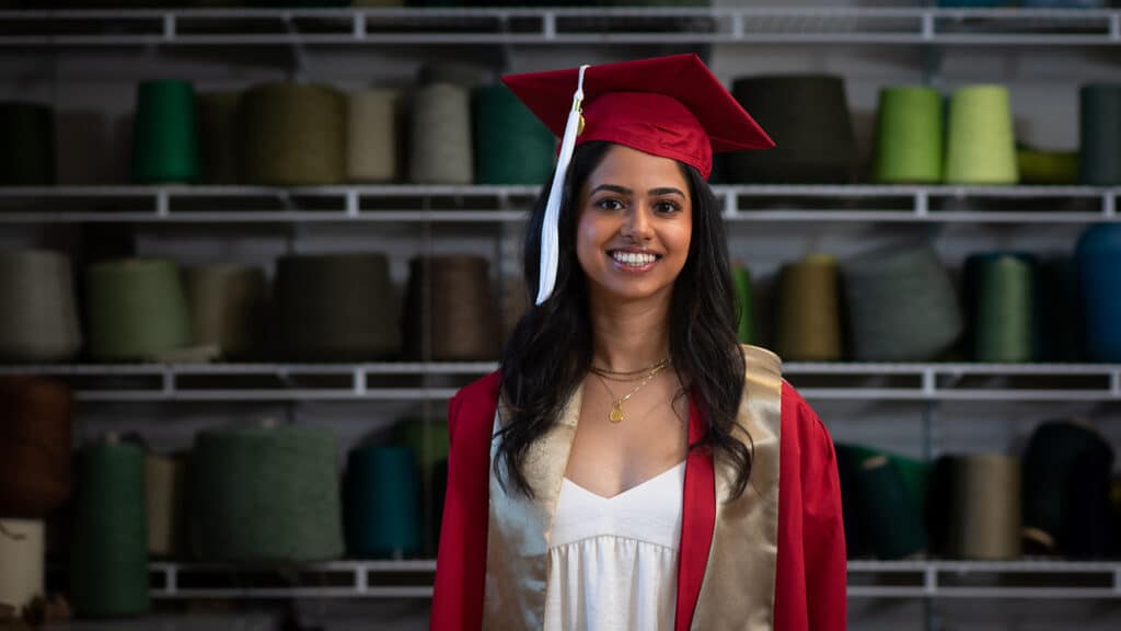 Ritika Shamdasani poses in front of shelves full of spools of yarn in a Wilson College of Textiles classroom wearing her cap and gown.