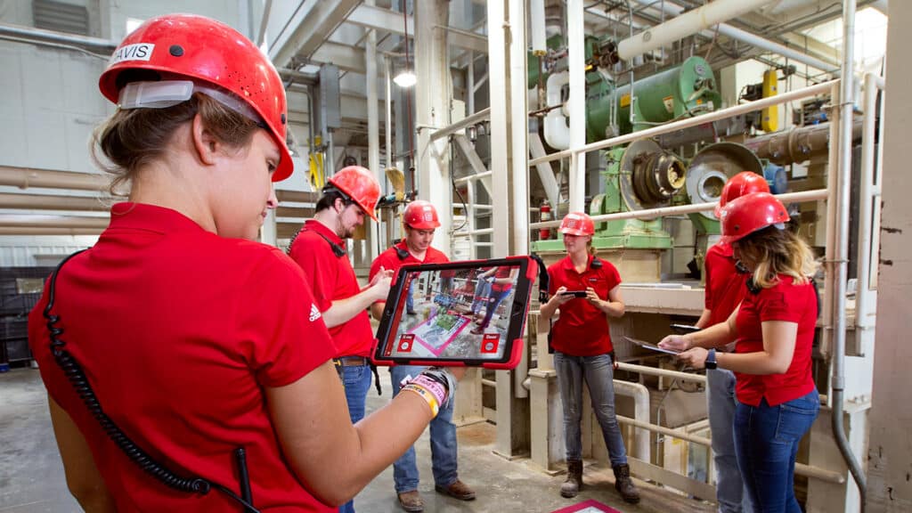 Undergraduate students in red shirts and hard hats look at augmented reality information on a tablet.