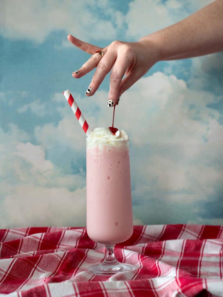 A hand with a cow-print manicure gently places a cherry on top of a cherry vanilla milkshake.