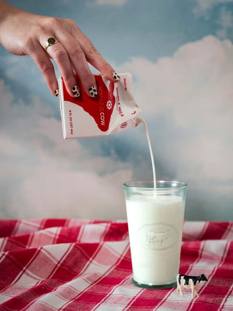 A hand with a cow-print manicure pours milk into a glass with a cow decal.