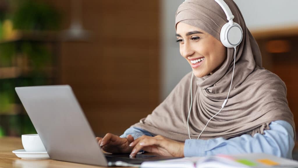 A woman wearing headphones works at her laptop, smiling.