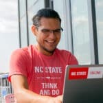 A man in an NC State T-shirt works at a laptop with a sticker reading "NC State Online."