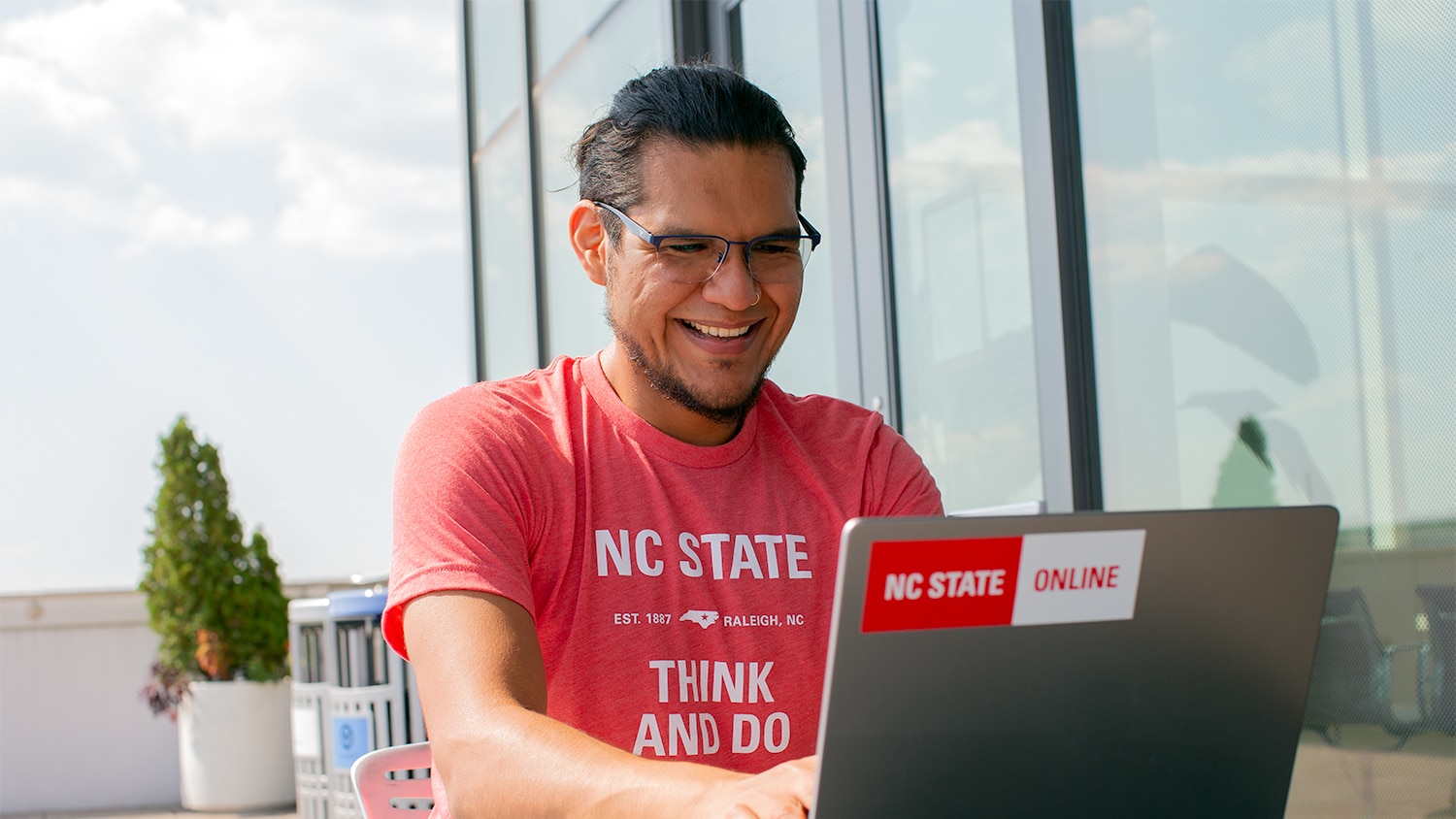 A man in an NC&#160;State T-shirt works at a laptop with a sticker reading "NC&#160;State Online."