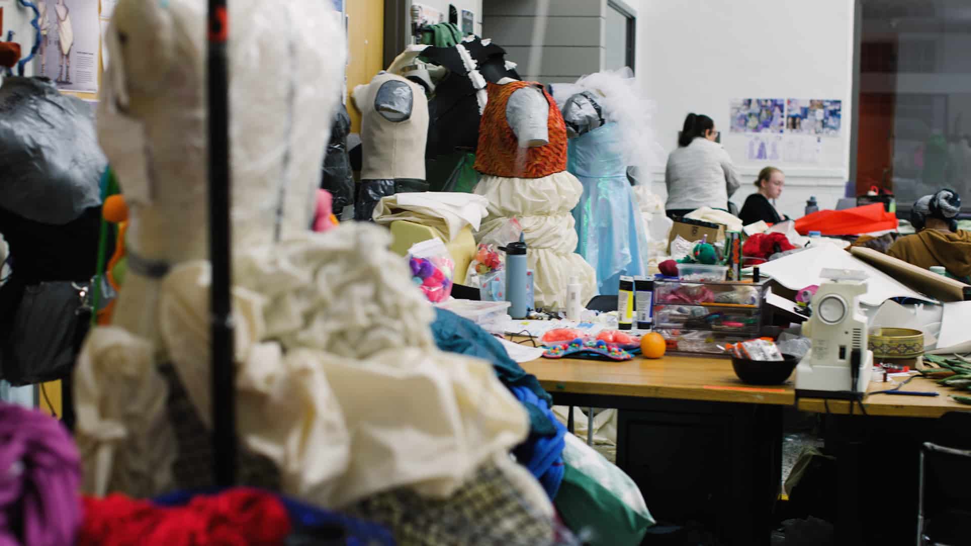 Dress forms with pieces of wearable art in varying stages of completion are arranged in a studio, with students working in the background on their collections for Art2Wear 2024.