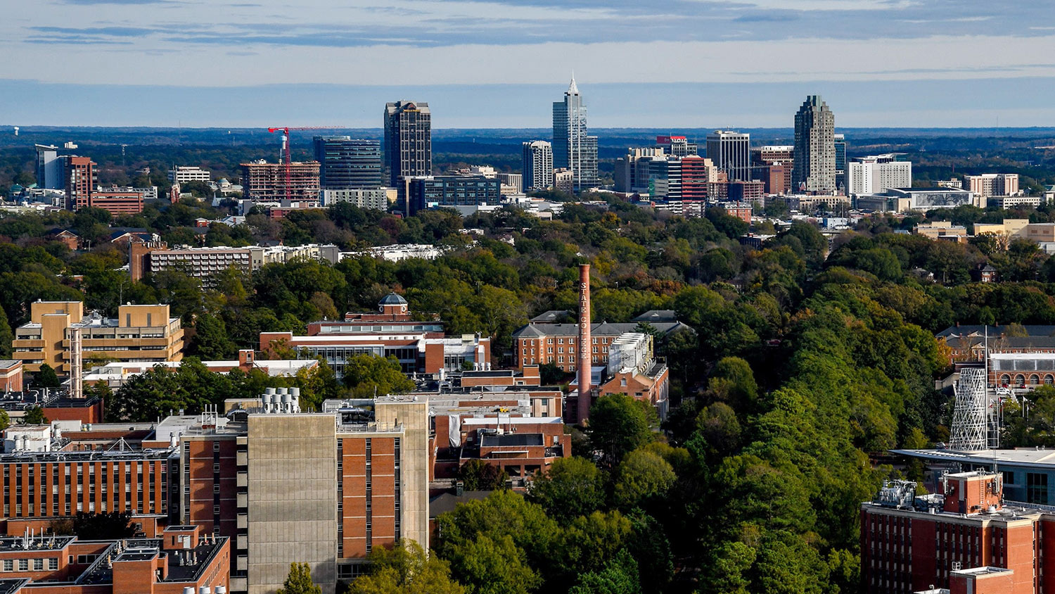 An aerial view of NC State's campus with the Downtown Raleigh skyline behind it.
