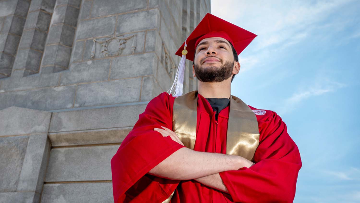 A brand-new NC State graduate stands with his arms crossed, donning his red cap and gown.