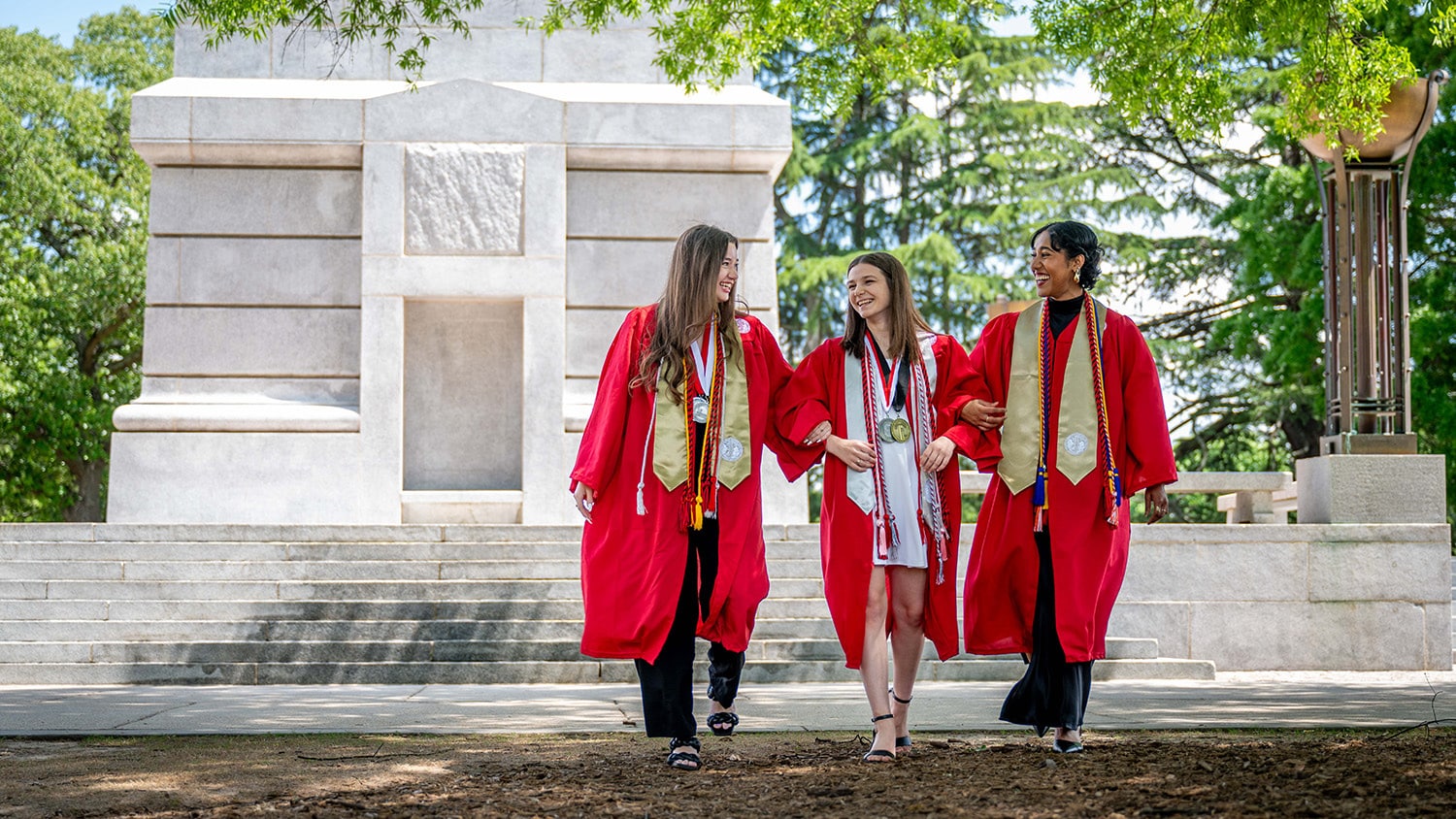 Three students in red graduation robes walk with arms linked outside of the Memorial Belltower.