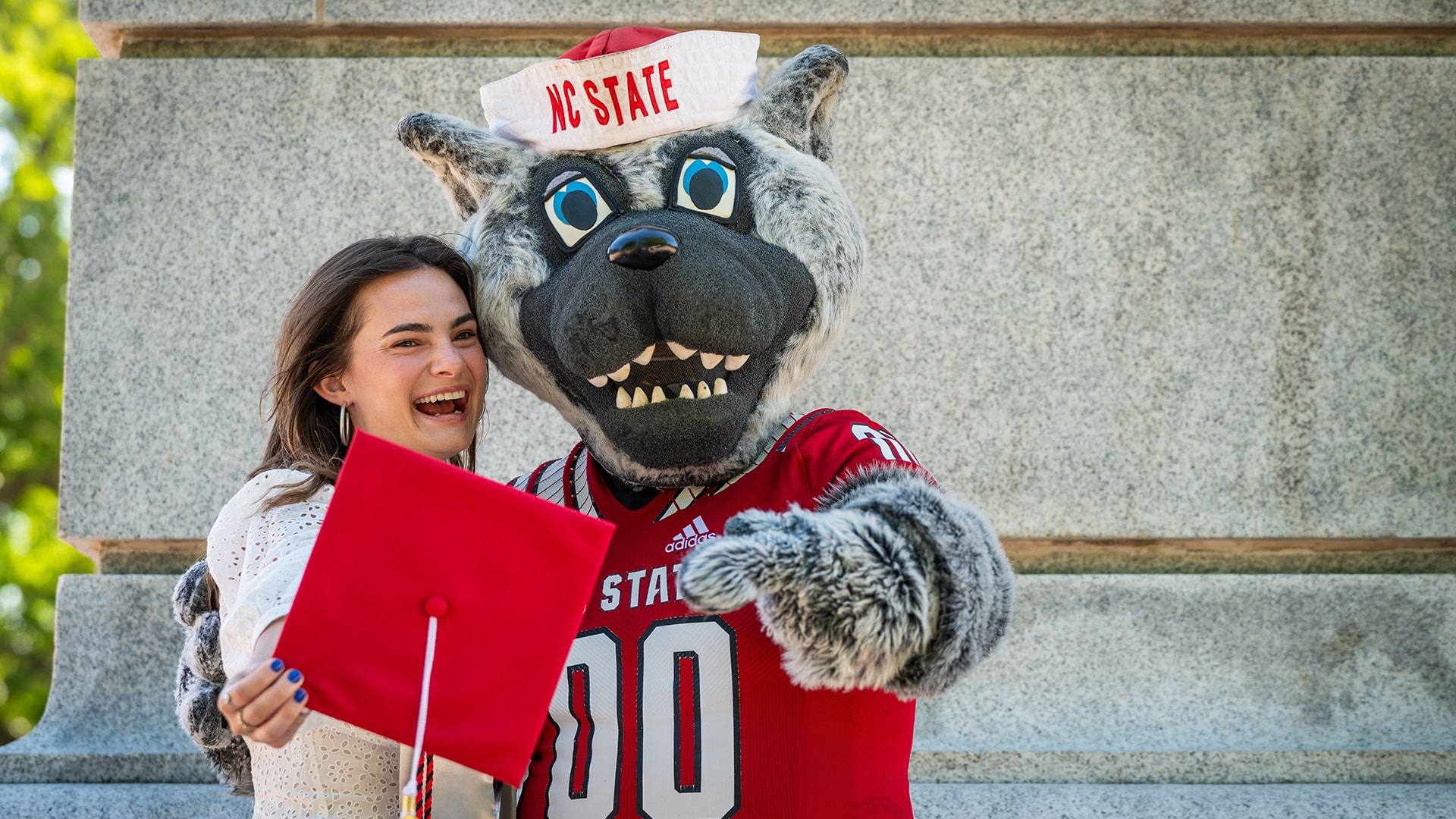 A student holds up her red graduation cap and poses with Mr. Wuf.