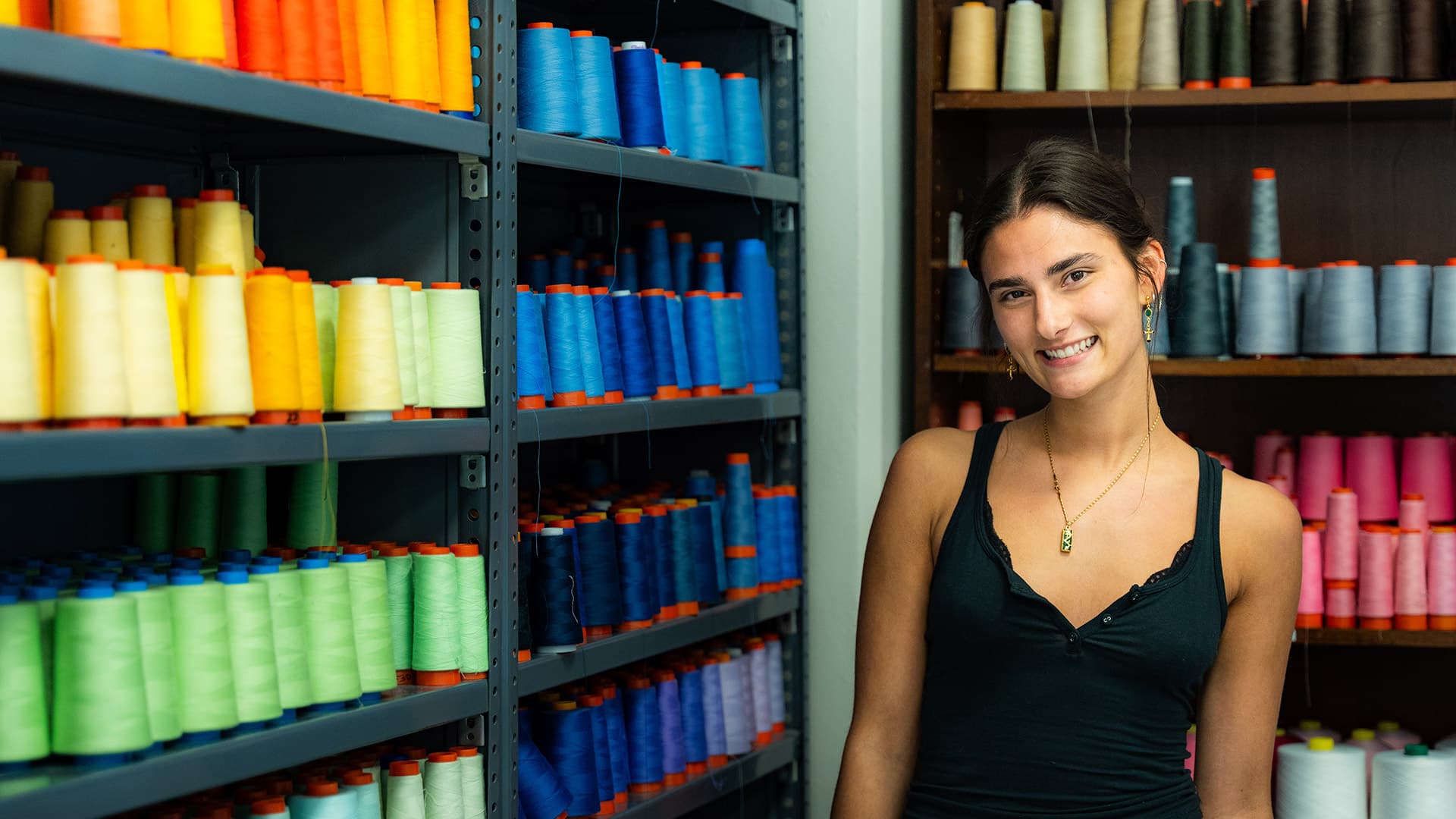 Fashion and textile design student Neely Mallik poses and smiles in a room filled with colorful spools of thread.