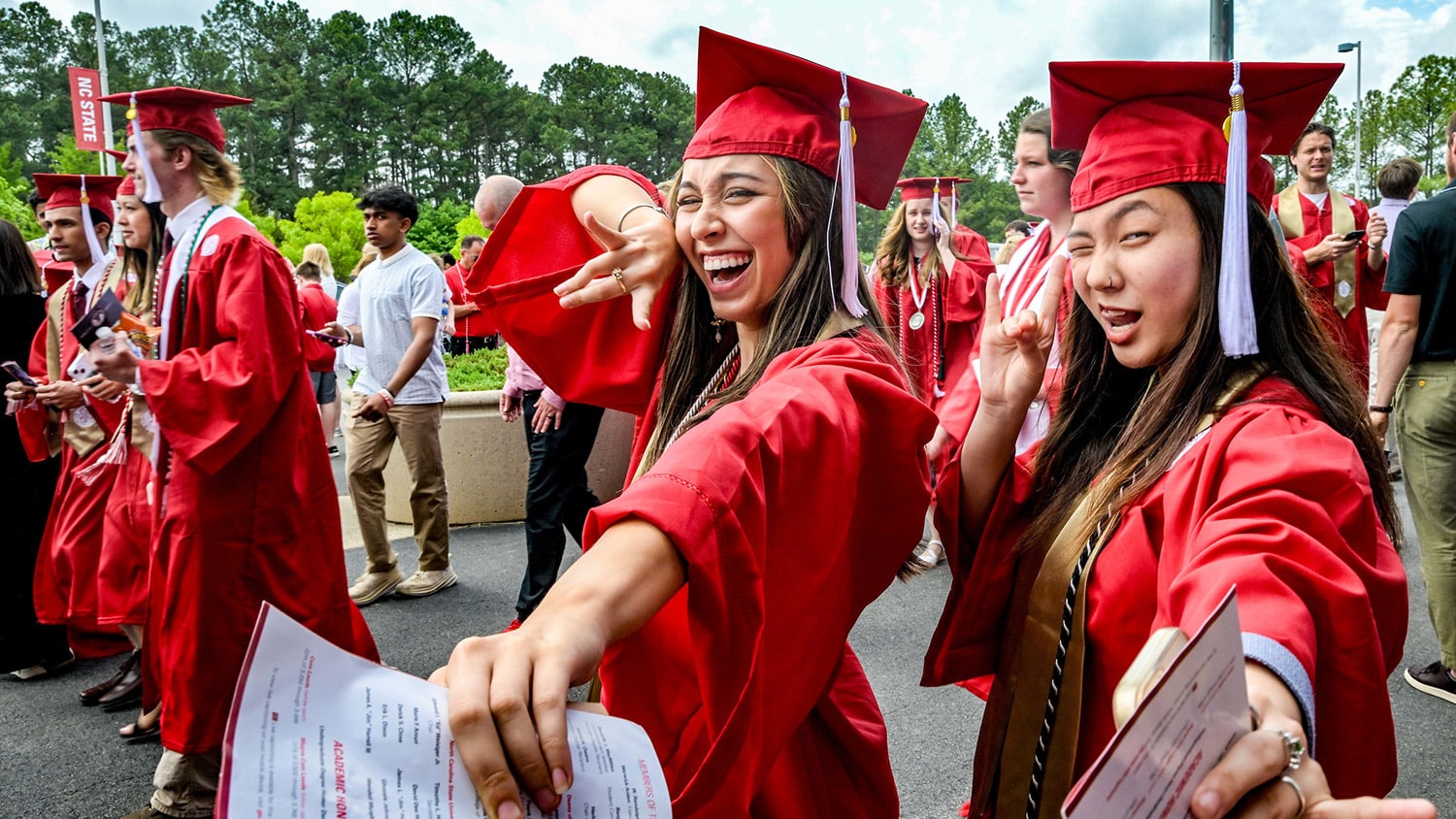 Two students in red graduation robes put up their wolfies and pose for a photo on their way to commencement.