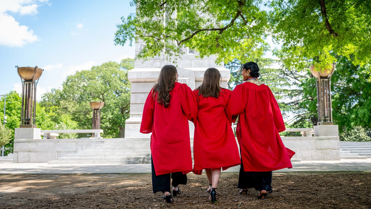Three new graduates in red robes walk toward the Memorial Belltower.