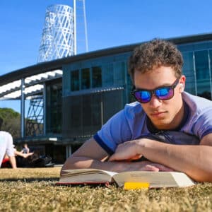 An NC State student dons shades to lay in the grass and read a book on a bright day outside Talley Student Union.