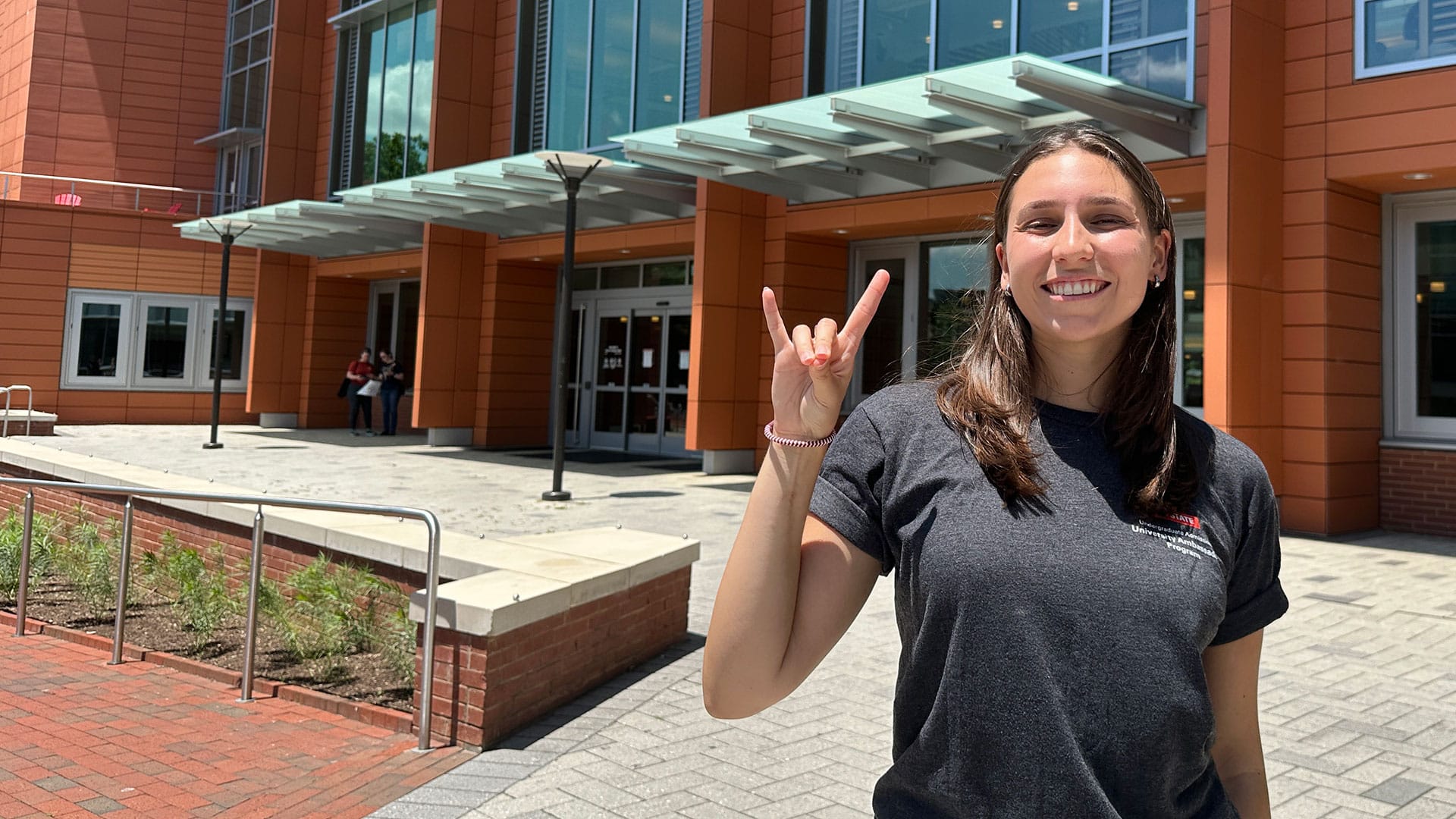 Abby Pfizenmaier, a rising fourth-year student at NC State and an Undergraduate Student Ambassador, smiles in front of the back entrance of Talley Student Union while showing the Wolf Ears hand signal.